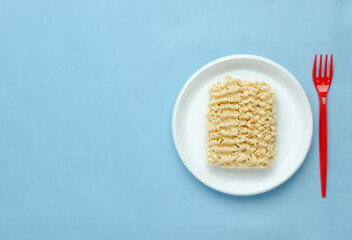 Top view of white plastic plate and raw noodels on it, red fork on the blue background.Empty space.Concept of plastic and unhealthy food