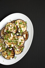 Homemade Grilled Eggplant with Feta and Herbs on a white plate on a black surface, top view. Flat lay, overhead, from above. Space for text.
