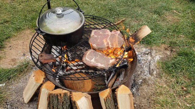 Grilling beef steaks over fire pit next to cost iron pot of boiling soup.