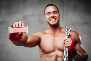 Handsome bodybuilder with chains and bandaged arms showing his favourite protein powerbar which he...