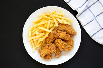 Homemade Crispy Chicken Wings and French Fries on a white plate on a black background, top view. From above, overhead, flat lay.