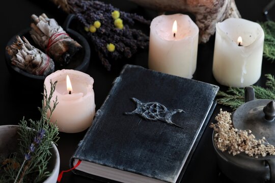 Wiccan witch altar with a hand made old looking book grimoire with triple moon symbol, and other various items - dried flowers, nature elements, burning candles, sage smudge sticks in black background
