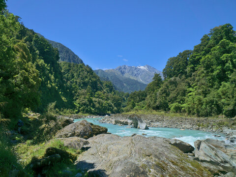 Lonely hiker on the Copland Track between the turquoise Copland River running through rocks, thick forest and snowy mountain peak, South Island, New Zealand