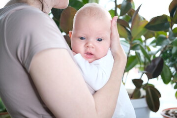 A beautiful young mother holds her newborn son in her arms by the window. Close-up portrait of a...