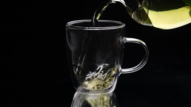 Green tea is being poured from the glass teapot ona black background