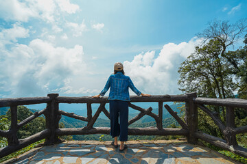 A photo from the back of a woman standing on an old grunge cement terrace of a high hill viewpoint. The scenery in front of her has mountains with lots of lush greenery and the sky on a cloudy day.