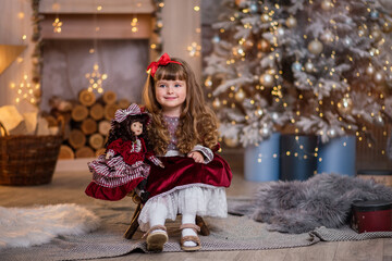 Little beautiful girl in a red dress near the Christmas tree. Girl with a doll are playing Christmas. The child smiles. Beautiful girl with long curly hair. child in santa claus clothes