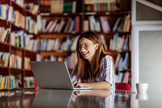 Young smiling attractive college girl sitting in library and using laptop. She is writing homework.