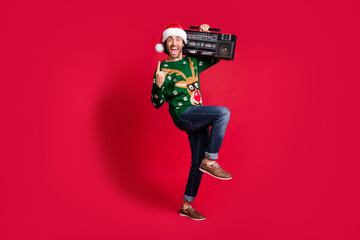 Full length body size view of his he attractive glad cheerful cheery guy wearing Santa hat dancing carrying boombox having fun show horn sign isolated bright vivid shine vibrant red color background