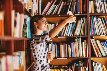 Young beautiful brunette in dress and with eyeglasses holding a book and taking selfie while...