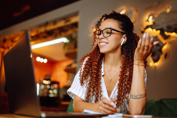 Portrait of African American woman sitting at cafe having video call on laptop computer. Woman, student wearing headphones calling on laptop, talk by webcam  with friend or relative, online course.