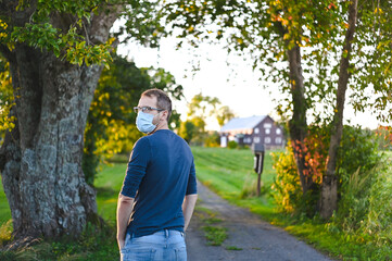 Naklejka na ściany i meble portrait of attractive nerd in long sleeve glasses and protective face mask outside in the park at sunset