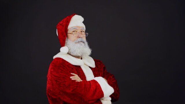Waist up portrait of Santa Claus in traditional costume crossing arms and nodding head confidently in slow motion while looking at camera standing against black background
