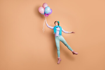 Fototapeta na wymiar Full body photo of pretty shocked lady jump high hold many air balloons raise up with wind blow wear violet jumper green pants boots blue beret scarf isolated beige color background