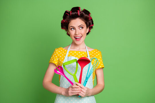 Photo of positive girl curious look copyspace hold spatula whisk spoon fork potato masher wear dotted yellow t-shirt hair rollers isolated over green color background