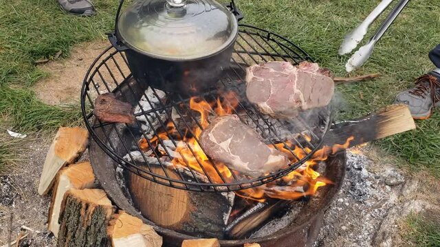 Beef steaks being checked and flipped while grilled over fire pit next to cast iron pot of boiling soup while at camp site.
