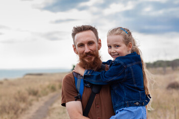 Father with daughter in hands standing on a hill with nature lanscape. Little blonde girl smiling and hugging with dad outdoor. Loving child embrace her daddy. Father's Day family greeting card.