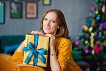 Obraz na płótnie Canvas A red-haired woman in a yellow sweater holds a gift wrapped in gold against the background of a Christmas tree.