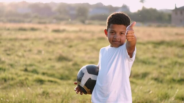 African boy is having fun playing alone in a summer field. The boy shows thumb up and playing football on sunset. The little kid is happy on holidays.