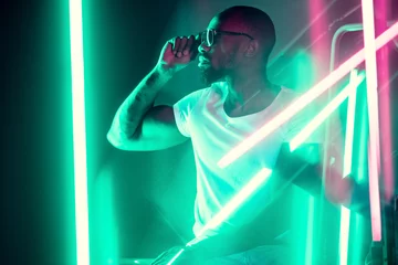 Schilderijen op glas Geometric lines. Cinematic portrait of stylish young man in neon lighted room. Bright neoned colors. African-american model, musician indoors. Youth culture in party, festival style and music concept. © master1305