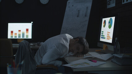 Tired businessman sleeping on work table front computer in dark office.