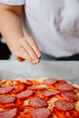 Close-up of a hand pouring pepper and herbs spices into a pepperoni pizza. Preparing homemade food for meeting friends.