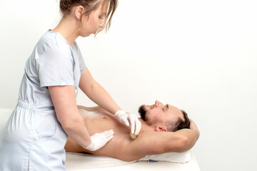 Young man receiving waxing underarm or epilation armpit by young female cosmetologist in beauty...