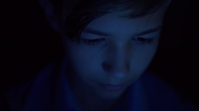 reflection of a bright multi-colored screen on the boy's face in complete darkness