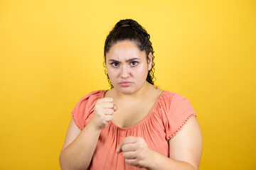 Young beautiful woman with curly hair over isolated yellow background Punching fist to fight, aggressive and angry attack, threat and violence