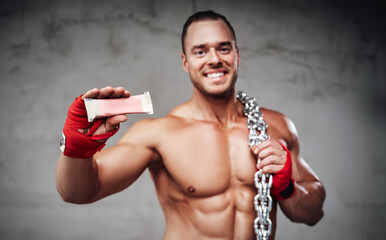 Handsome bodybuilder with chains and bandaged arms showing his favourite protein powerbar which he...