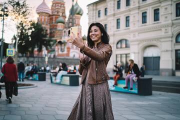 Fototapeta premium Cheerful female travel blogger shooting influence vlog with web sightseeing during solo vacations in Moscow, smiling Asian tourist in earbuds making international video call for communicate in roaming