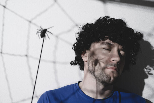 Portrait of an adult man in a black wig with a shadow of a spider and cobwebs on his face. Creates creepy puppet shadows. Halloween. Copy space