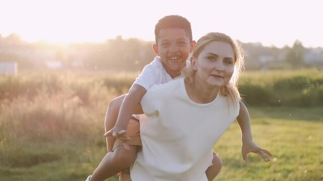 A happy mother is having fun with her son. The kid is at mother's back smiling. Sunset. Happy family concept.
