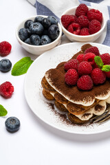 portion of Classic tiramisu dessert with raspberries and blueberries on ceramic plate isolated on white