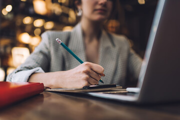 Cropped image of intelligent writer using laptop computer for elearning and informative research during education exam preparation, selective focus on female hand with pencil studying at table desk