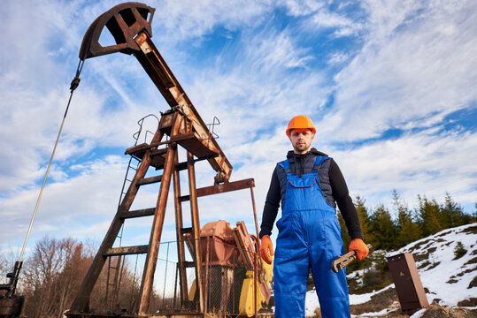 Portrait of oil well enginner in protective helmet and work overalls holding industrial wrench. Male worker standing near petroleum pump jack under beautiful sky. Concept of petroleum industry.