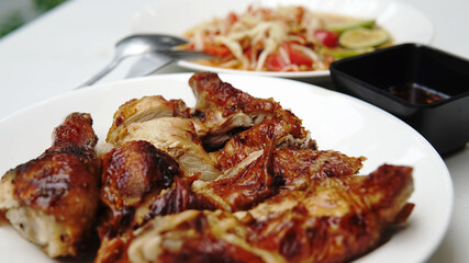 Thai food, chicken roasted or grilled on white plate with blurred dipping sauce cup and papaya salad on white background. selective focus