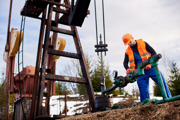 Oil worker in blue overalls, orange vest and helmet using industrial wrench while repairing pipe of...
