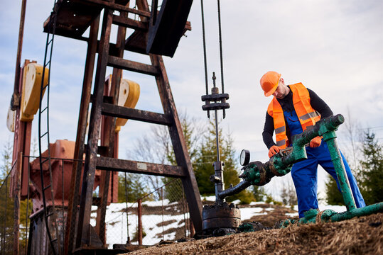 Oil Worker In Blue Overalls, Orange Vest And Helmet Using Industrial Wrench While Repairing Pipe Of Oil Well Pump Jack, Tightening Bolt On Pipe. Concept Of Petroleum Industry And Repair.