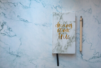 white marble notebook with text dreams come true  and beige pen on white marble background. Stationery flatlay.