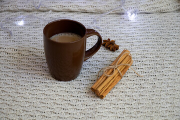 A brown cup of coffee, next to cinnamon sticks and star anise. Knitted background and Christmas lights. Back.