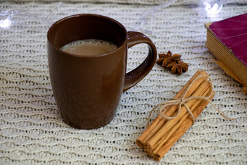 A brown cup of coffee, next to cinnamon sticks and star anise. Knitted background and Christmas lights. Back.