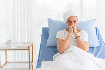 Front view of senior breast cancer patient wearing a headscarf laying in bed and praying in the hospital room, cancer concept