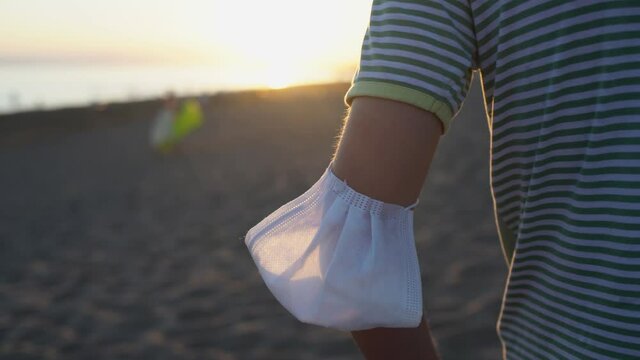 Closeup back view of young kid wearing medical mask on arm standing alone on sunset summer sandy beach enjoying fresh air.