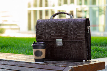 Dark brown leather men's briefcase and a cup of coffee on wooden bench outdoor. Sunny afternoon in a park