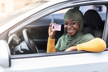 Happy black muslim woman in hijab sitting in car, showing driver license - obrazy, fototapety, plakaty