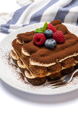 portion of Classic tiramisu dessert with raspberries and blueberries on ceramic plate isolated on white background with clipping path