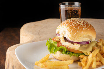 Hamburger and French fries placed on a white plate together with a glass of soft drink on rustic wood with black background, selective focus.