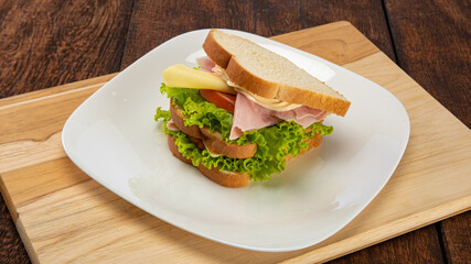 Large loaf bread sandwich, mayonnaise, cheese, ham, lettuce and tomato on a white plate on wood on a rustic table, selective focus.