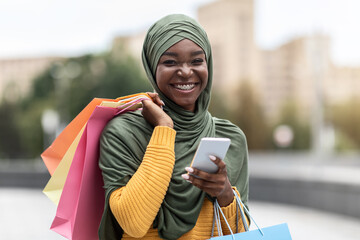 Discount App. Black muslim woman standing with smartphone and shopping bags outdoors